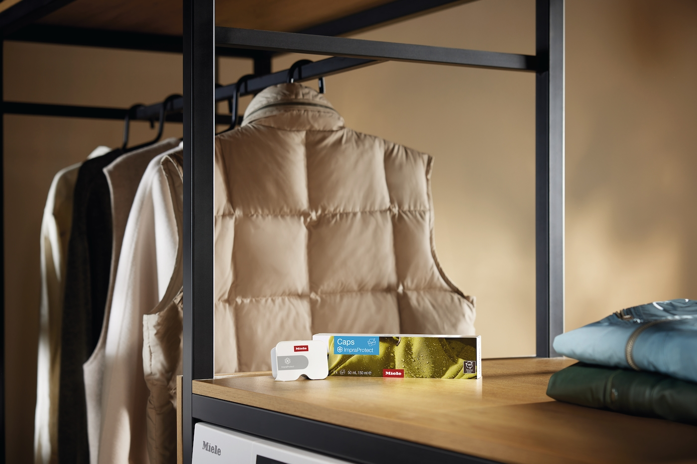 Ein Steppweste hängt neben Kleidung. Miele ImpradProtect Caps sind auf einem Regal.