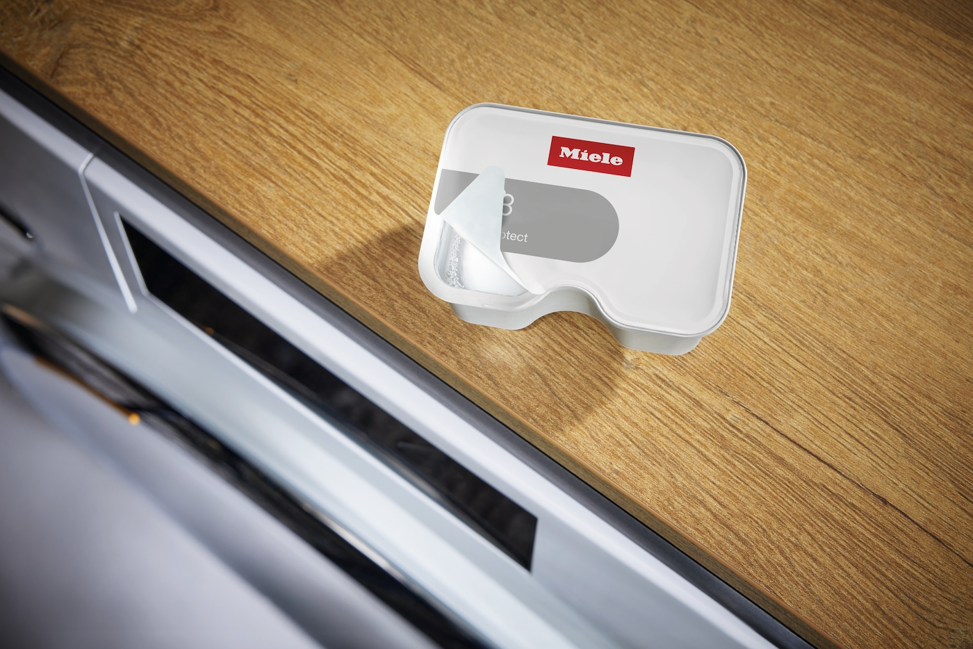 Ein Miele Reiniger-Tab liegt auf einem Holztisch neben einer Waschmaschine.