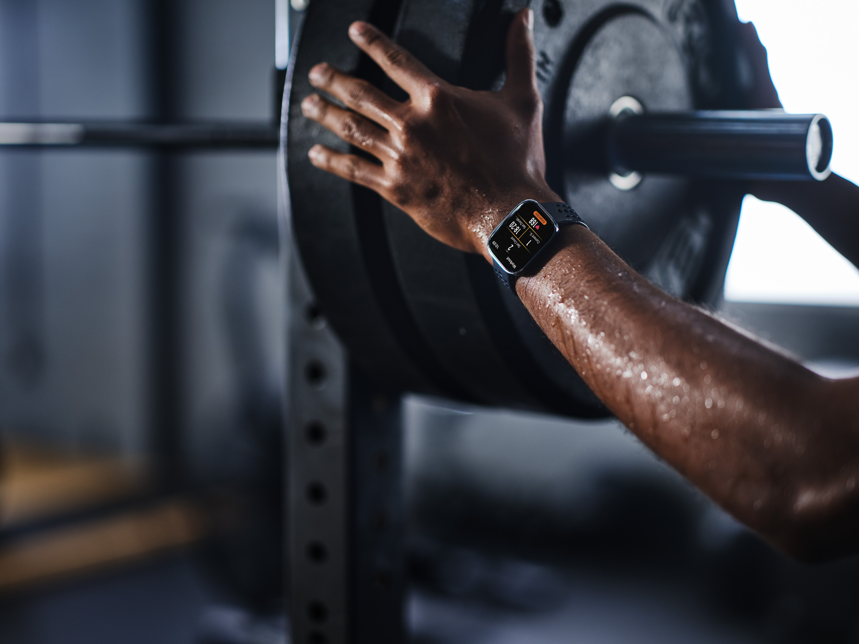 Spocona ręka kładzie talerz na sztandze. Smartwatch wyświetla dane treningu.