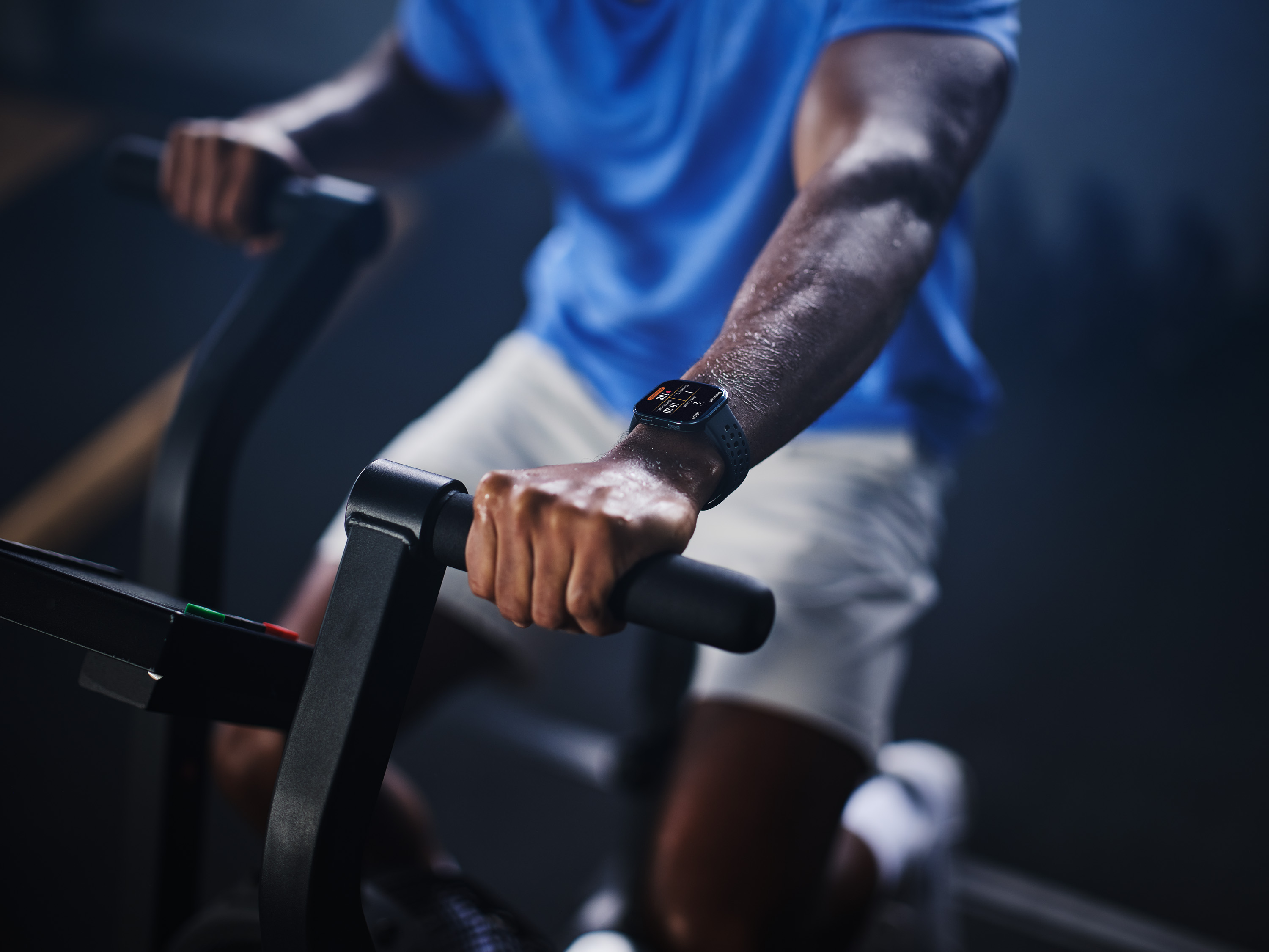 Mężczyzna w niebieskiej koszulce ćwiczy na rowerze stacjonarnym, nosi smartwatch.