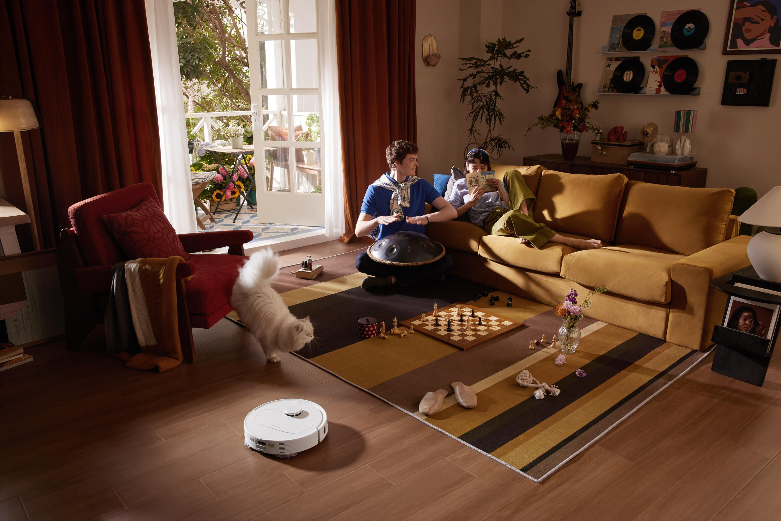 Ein weißer Kater läuft auf einem Holzboden, während zwei Personen im Wohnzimmer sitzen.