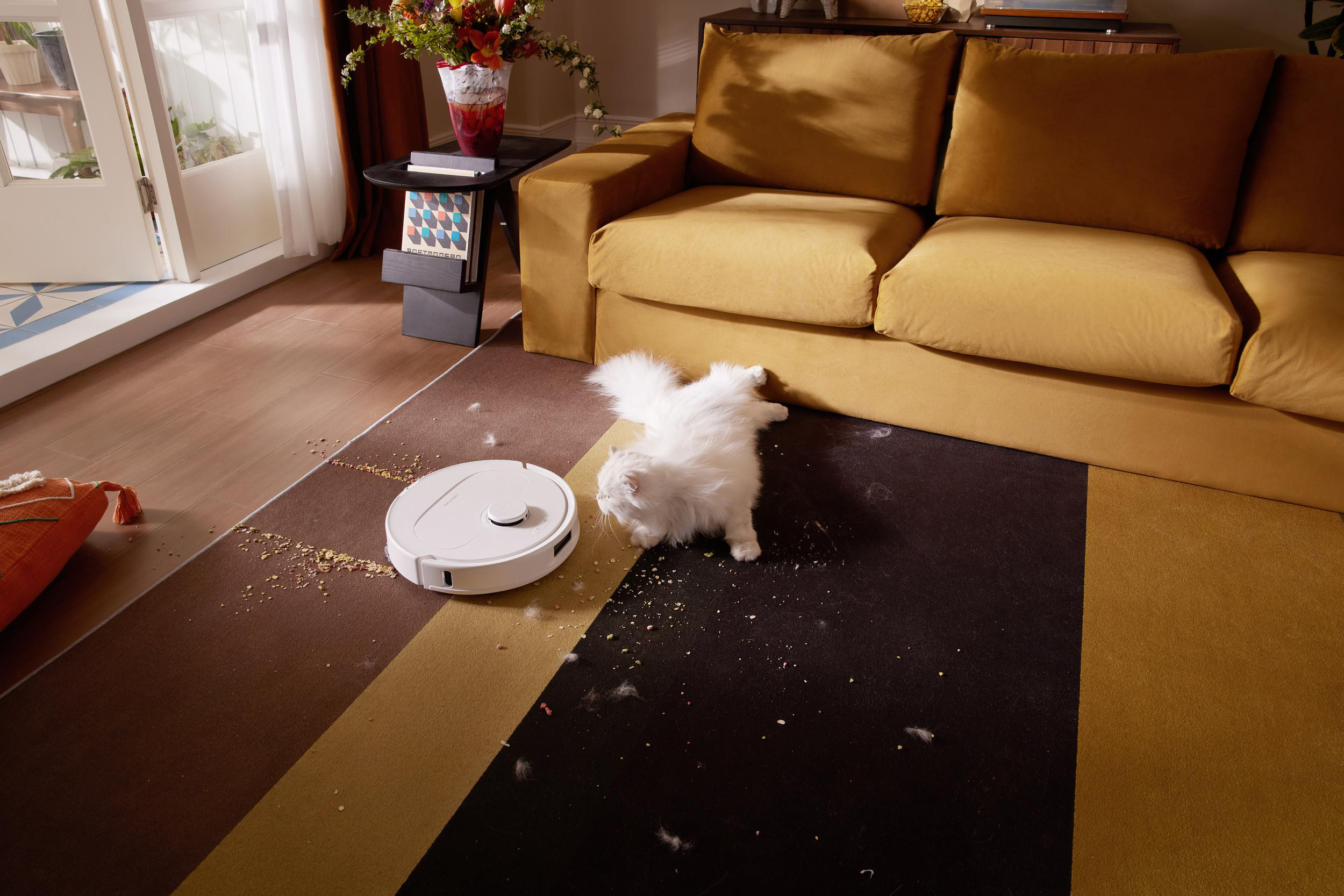 Ein weißer Saugroboter reinigt einen Teppich mit Essensresten und Haaren, während eine Katze zusieht.