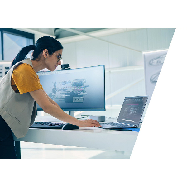 Una mujer con camisa amarilla y chaleco trabaja en una laptop y monitor grande con modelos 3D.