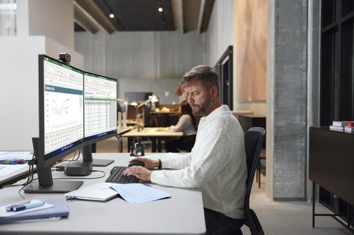 Hombre con suéter blanco trabajando en dos monitores en oficina moderna.