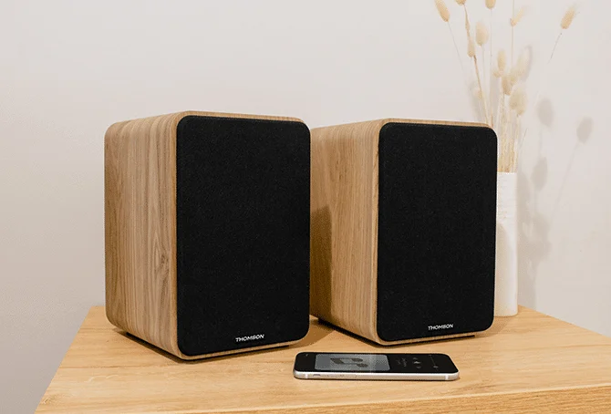 Dos altavoces de madera con frentes negros en una mesa, con un smartphone cerca.