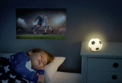 Mały chłopiec śpi w łóżku, lampka nocna w kształcie piłki nożnej świeci na szafce nocnej.