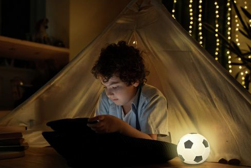 Chłopiec leży w namiocie, patrząc na tablet, obok niego lampka w kształcie piłki nożnej.