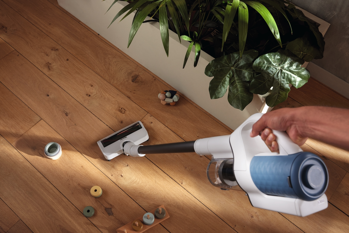 Hand hält weißen Akku-Staubsauger auf Holzboden mit Spielzeug und Pflanzen.