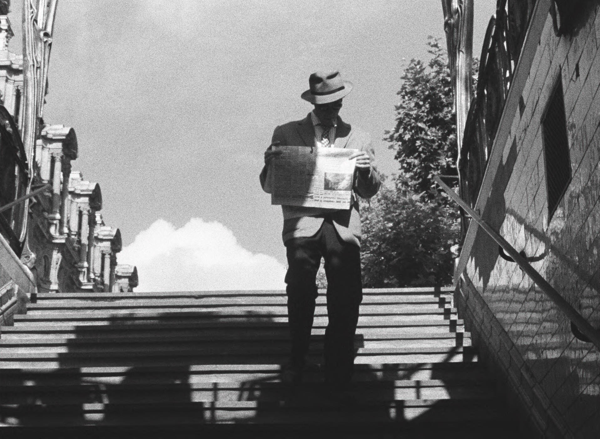 Mann mit Hut liest Zeitung auf Treppen. Gebäude und Bäume im Hintergrund.