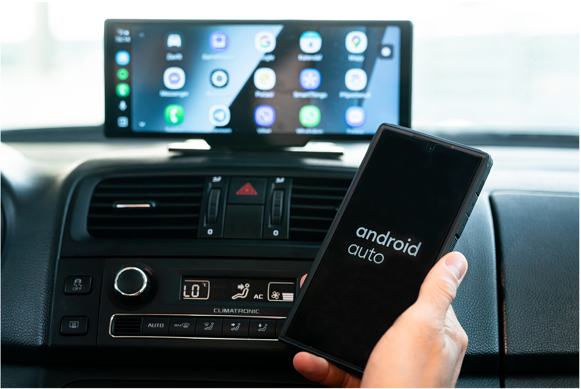 Kéz okostelefont tart az Android Auto felirattal a műszerfal előtt.