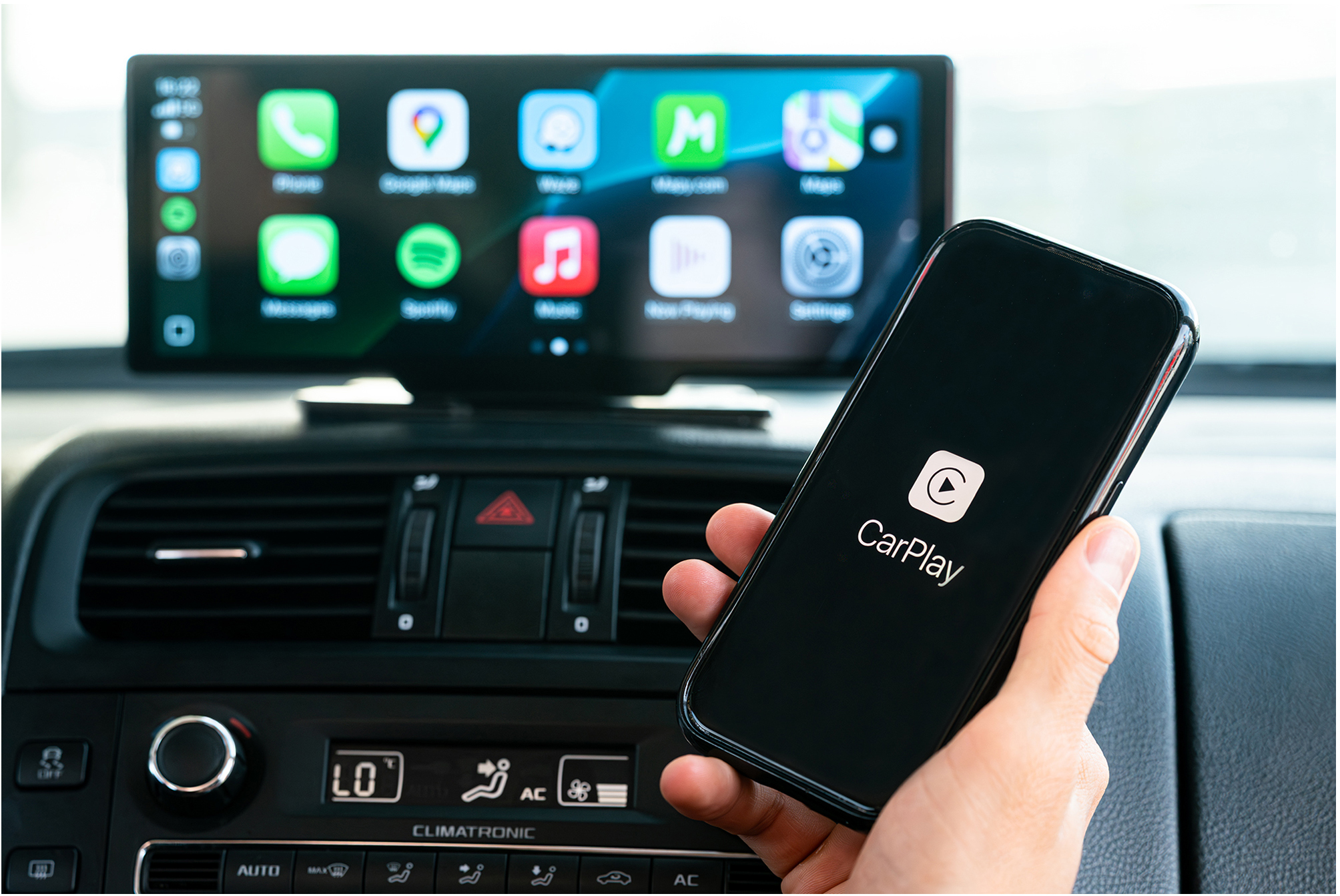Kézzel tartott okostelefon CarPlay logóval az autós rádió kijelzője előtt, app ikonokkal.