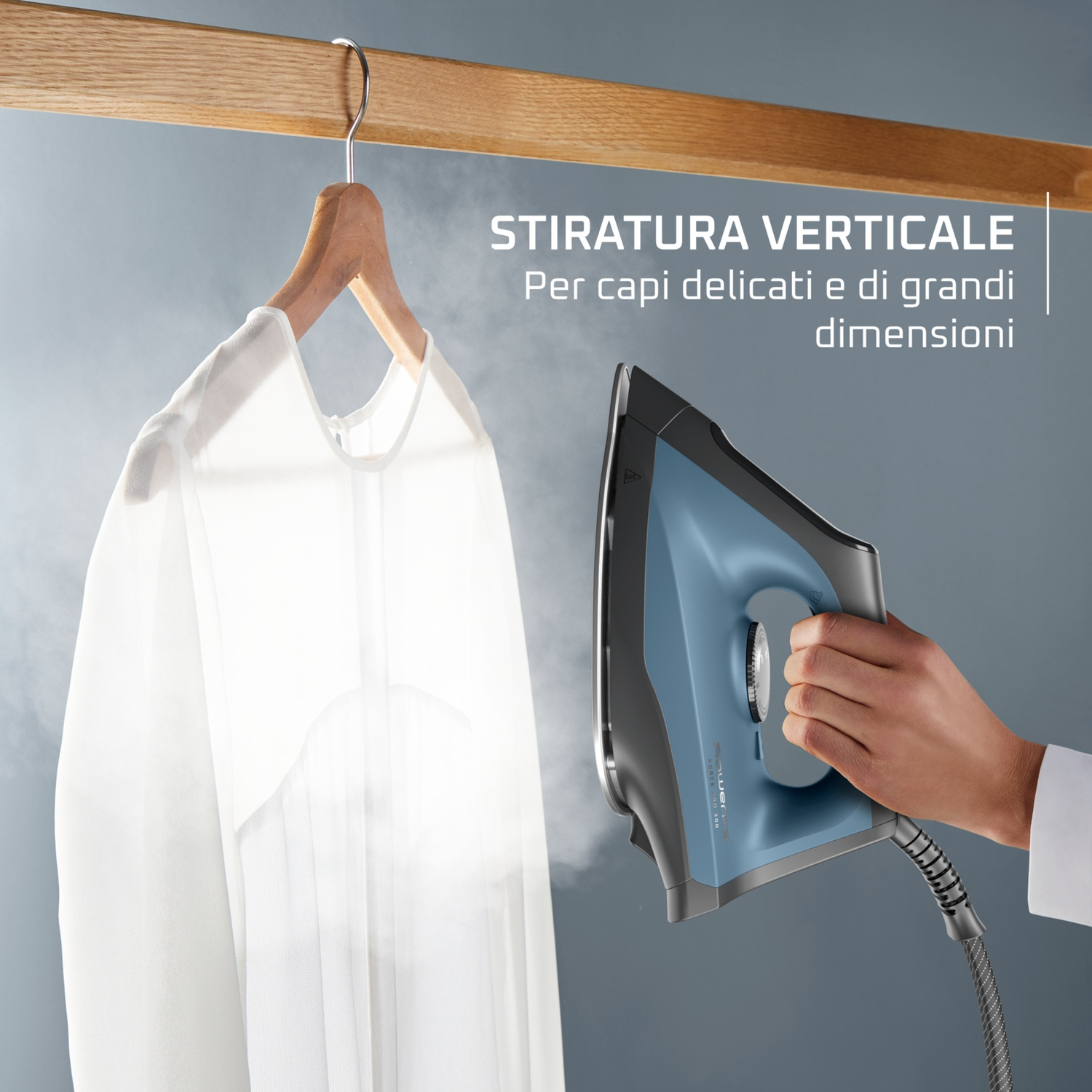 Una mano tiene un ferro da stiro blu, che spruzza vapore su una camicia bianca appesa a una gruccia di legno.
