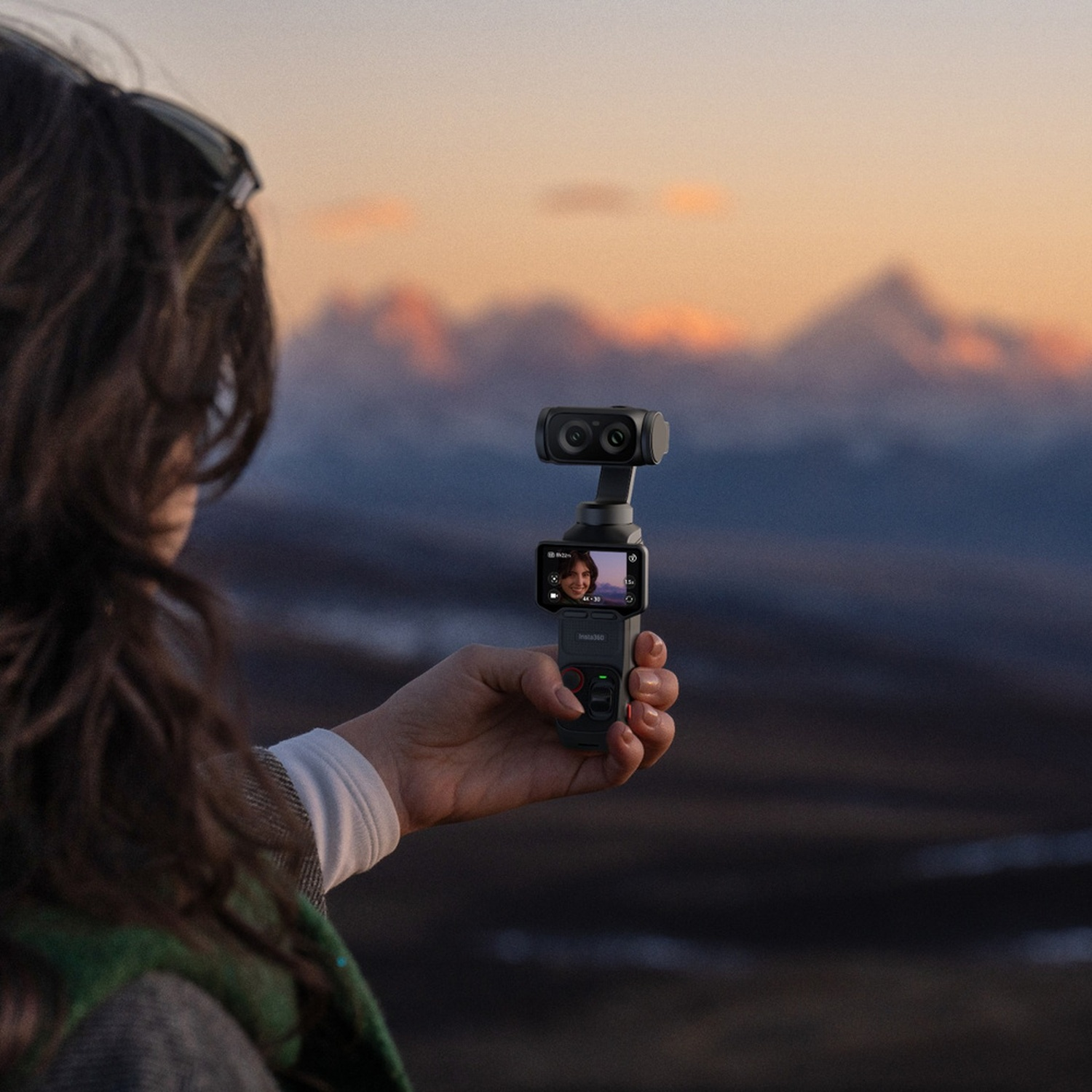 Una mano tiene una fotocamera con gimbal su un paesaggio montano al tramonto.