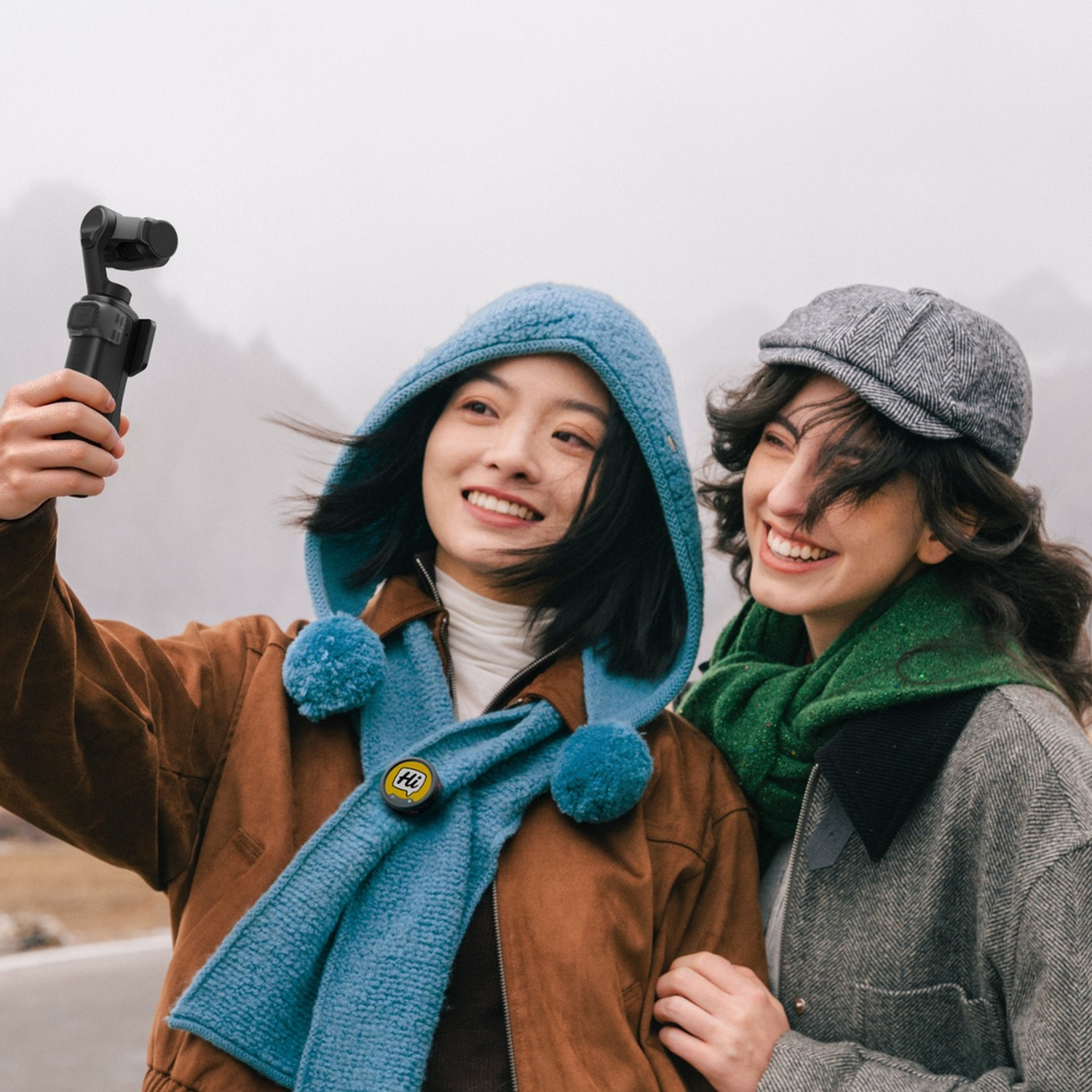 Due donne si scattano un selfie con un gimbal davanti a un paesaggio montano nebbioso.