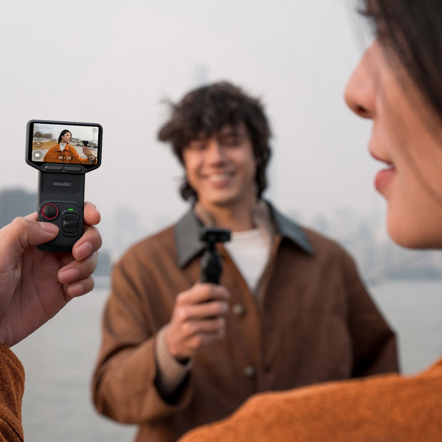 Una mano tiene una fotocamera con uno schermo che mostra una donna. Un uomo sorride sullo sfondo.