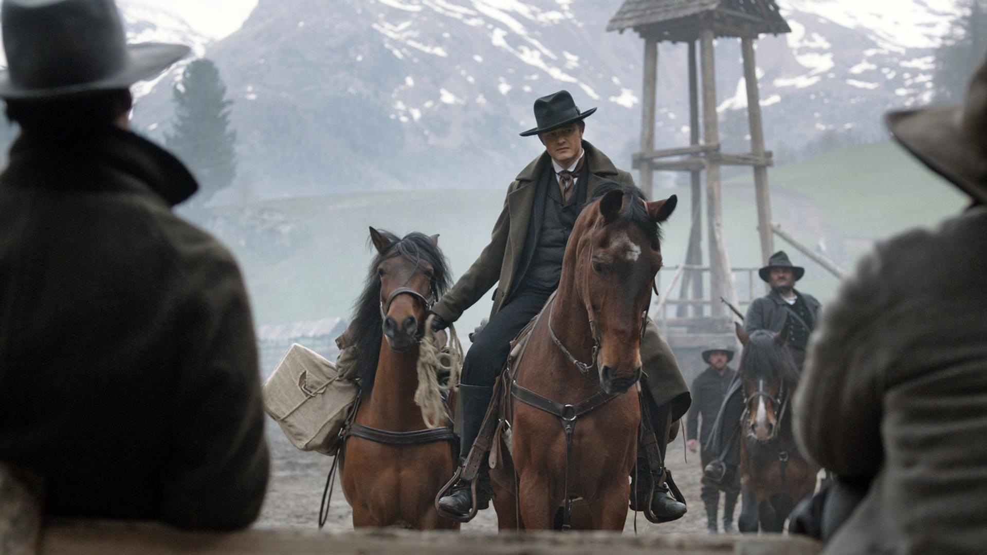 Männer in Hüten reiten auf Pferden vor Bergkulisse mit Holzturm.