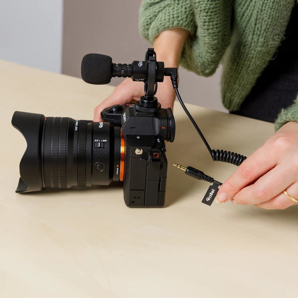 Eine schwarze Sony-Kamera mit Objektiv und Mikrofon steht auf einem Holztisch. Eine Hand hält ein Rollei-Kabel.