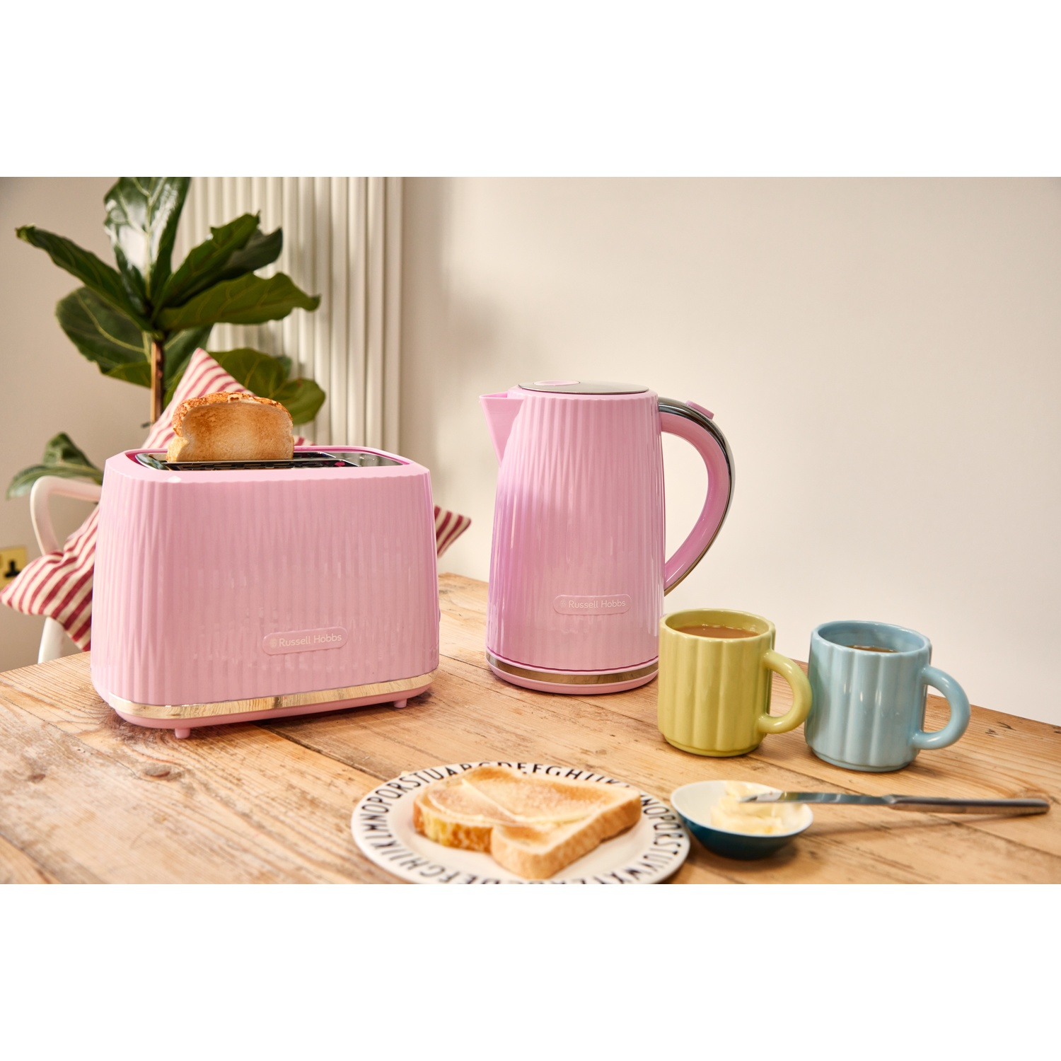 Een roze broodrooster met toast, een roze waterkoker en twee mokken op een houten tafel.