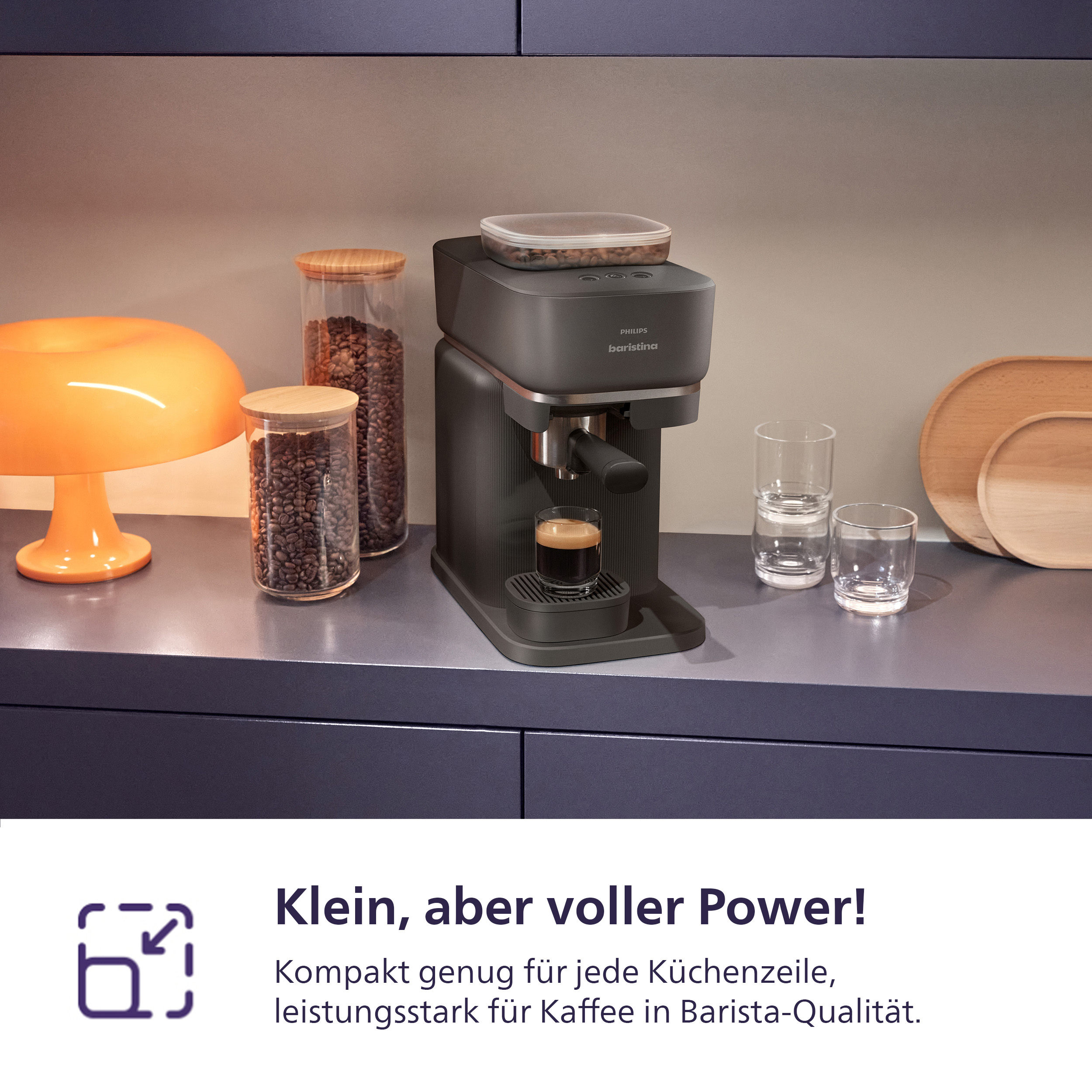 Dunkelgraue Kaffeemaschine mit Espresso in einem Glas, daneben Gläser und Kaffeebohnen.