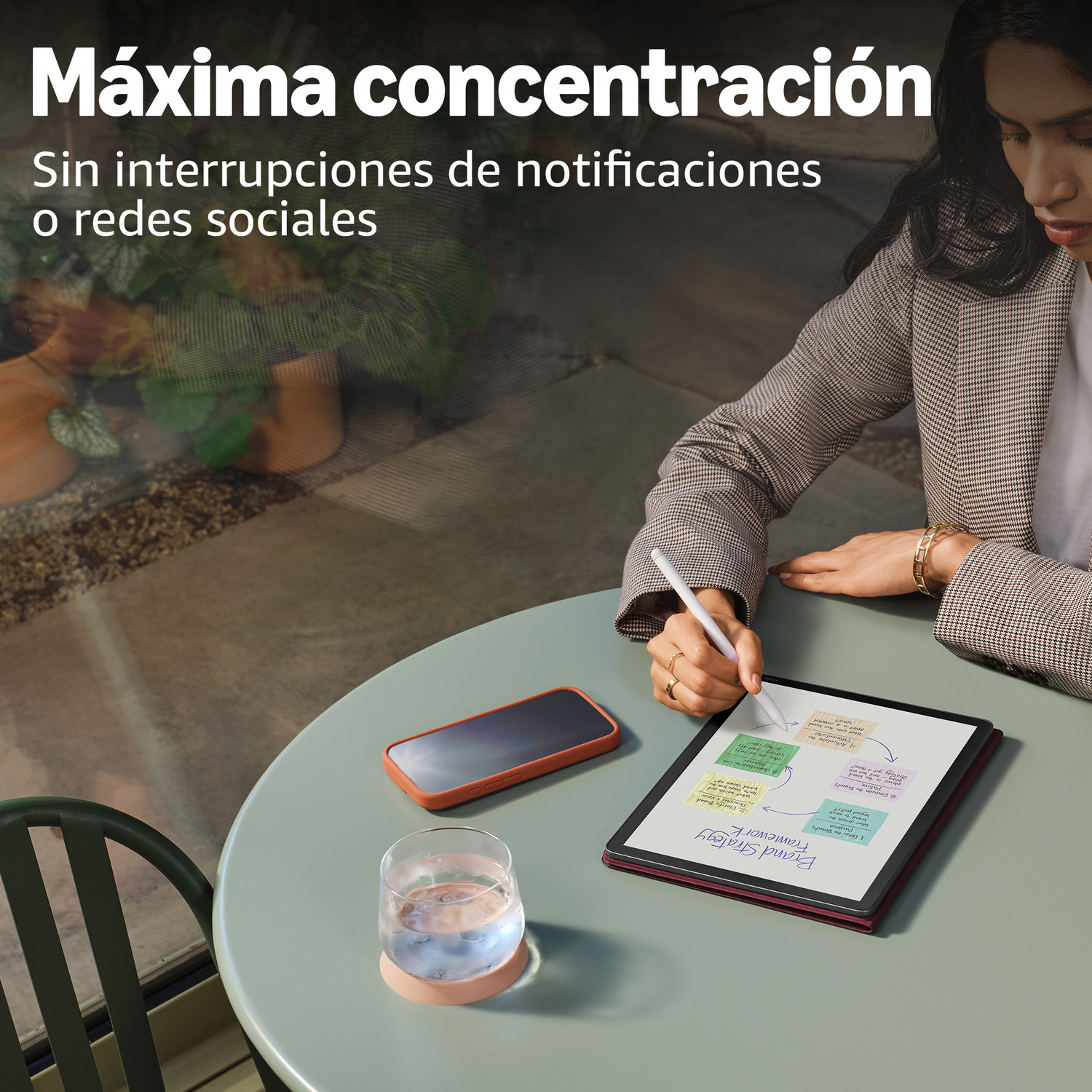 Mujer con lápiz óptico trabaja en tableta con notas. Teléfono y vaso de agua cerca.