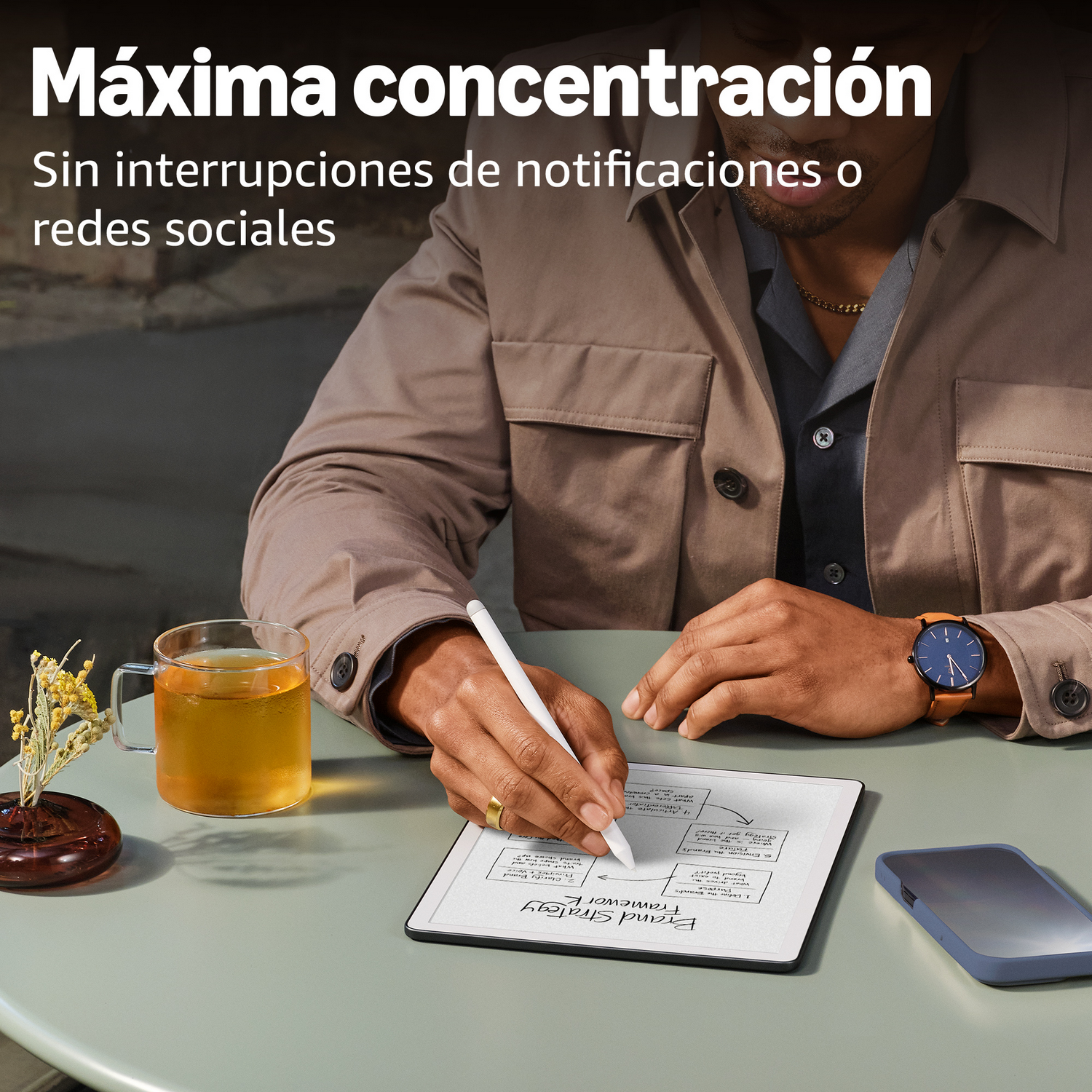 Hombre escribe en e-reader, té, teléfono y planta en mesa. Enfoque en concentración.