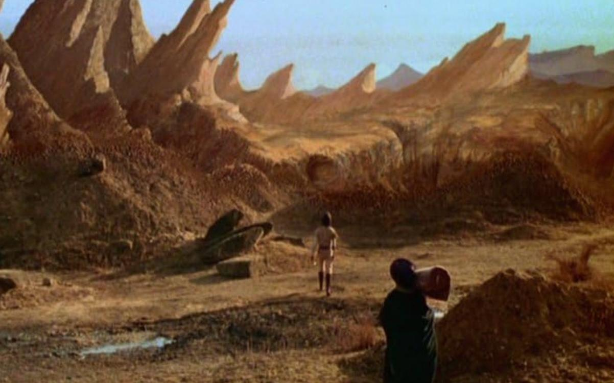 Zwei Figuren in einer trockenen, felsigen Landschaft mit spitz zulaufenden Felsformationen.