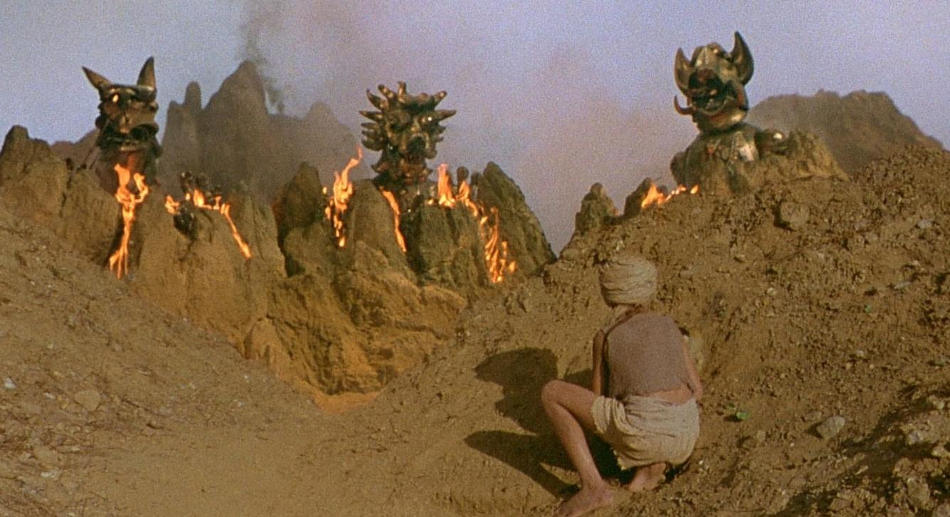 Ein Mann kniet in einer felsigen Landschaft mit drei grotesken goldenen Figuren, die auf Hügeln stehen und Feuer speien.