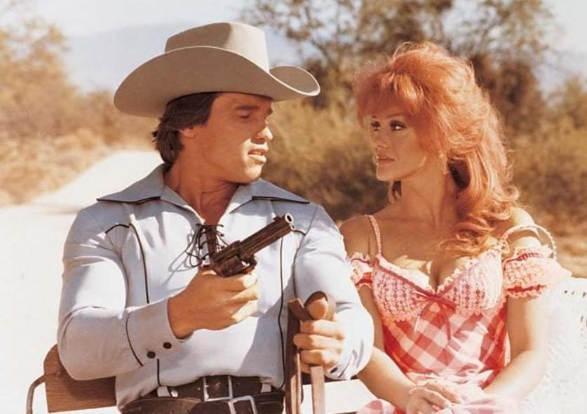 Ein Mann im Cowboyhut mit Waffe und eine Frau mit roten Haaren sitzen.