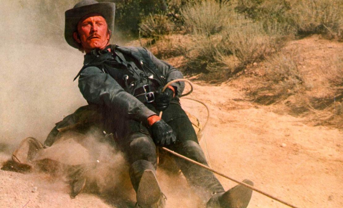 Ein Mann in einem Cowboyhut und dunkler Kleidung fällt im Staub auf einem felsigen Hang.