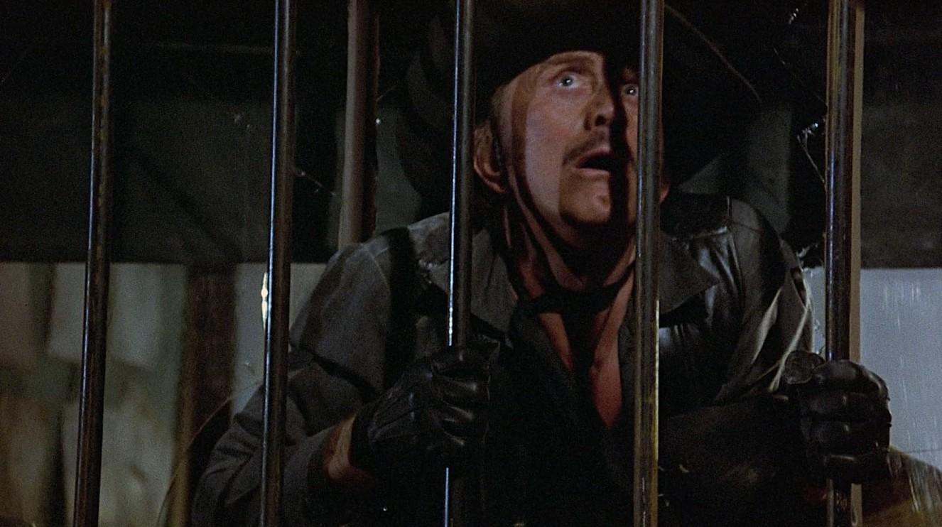 Ein Mann mit Hut und Ledermantel sitzt hinter Gittern und blickt besorgt nach oben.
