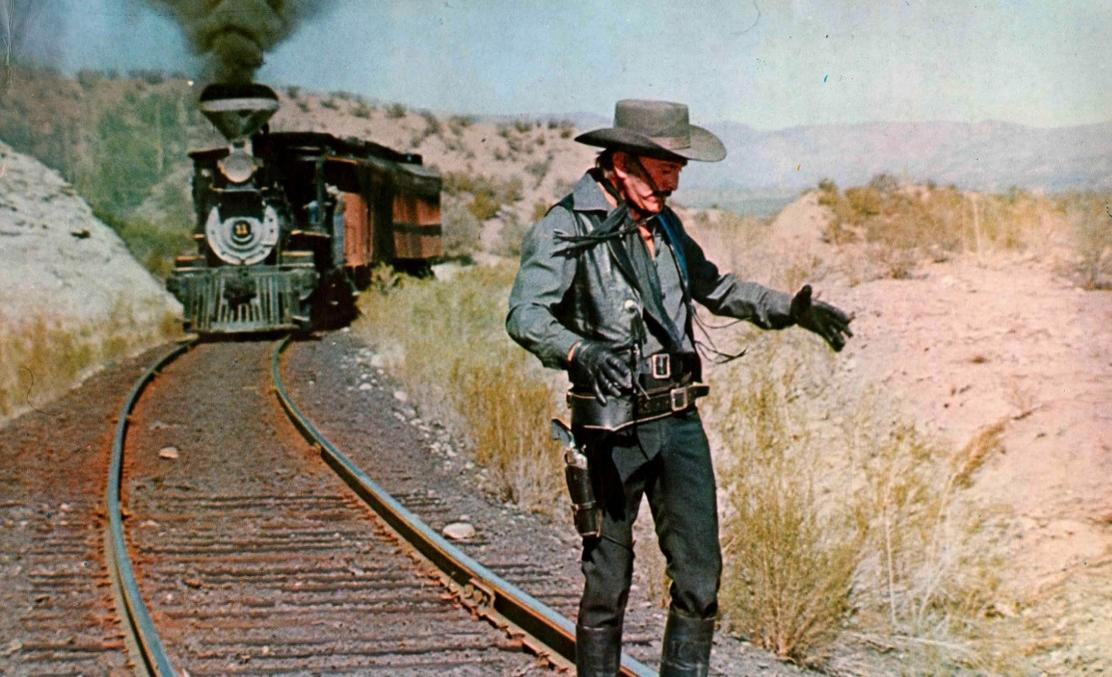 Ein Mann im Western-Look steht neben einer dampfenden Lokomotive in der Wüste.
