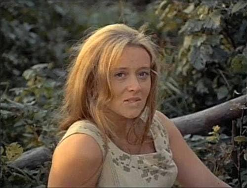Eine Frau mit blonden Haaren sitzt im Freien, umgeben von grüner Vegetation.