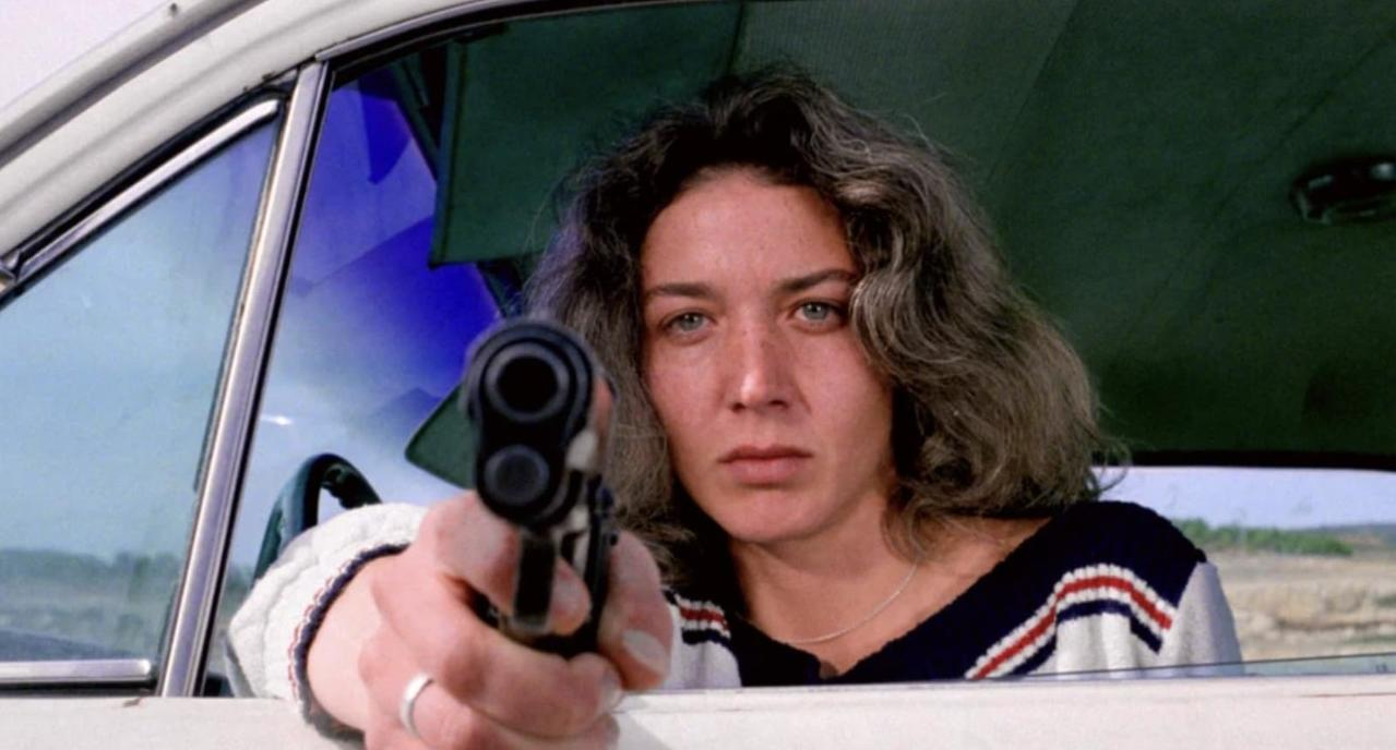 Eine Frau mit Locken zielt mit einer Pistole aus einem Auto auf den Betrachter.
