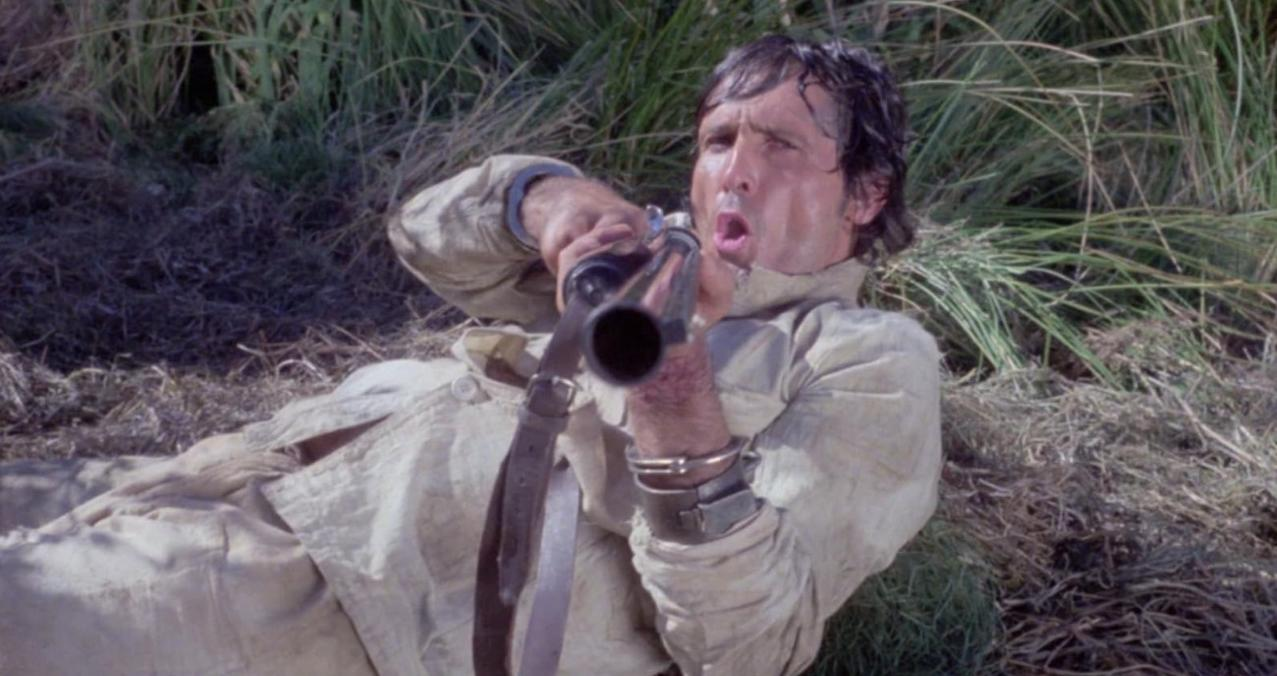 Mann in beige Kleidung liegt im Gras und zielt mit einem Gewehr.