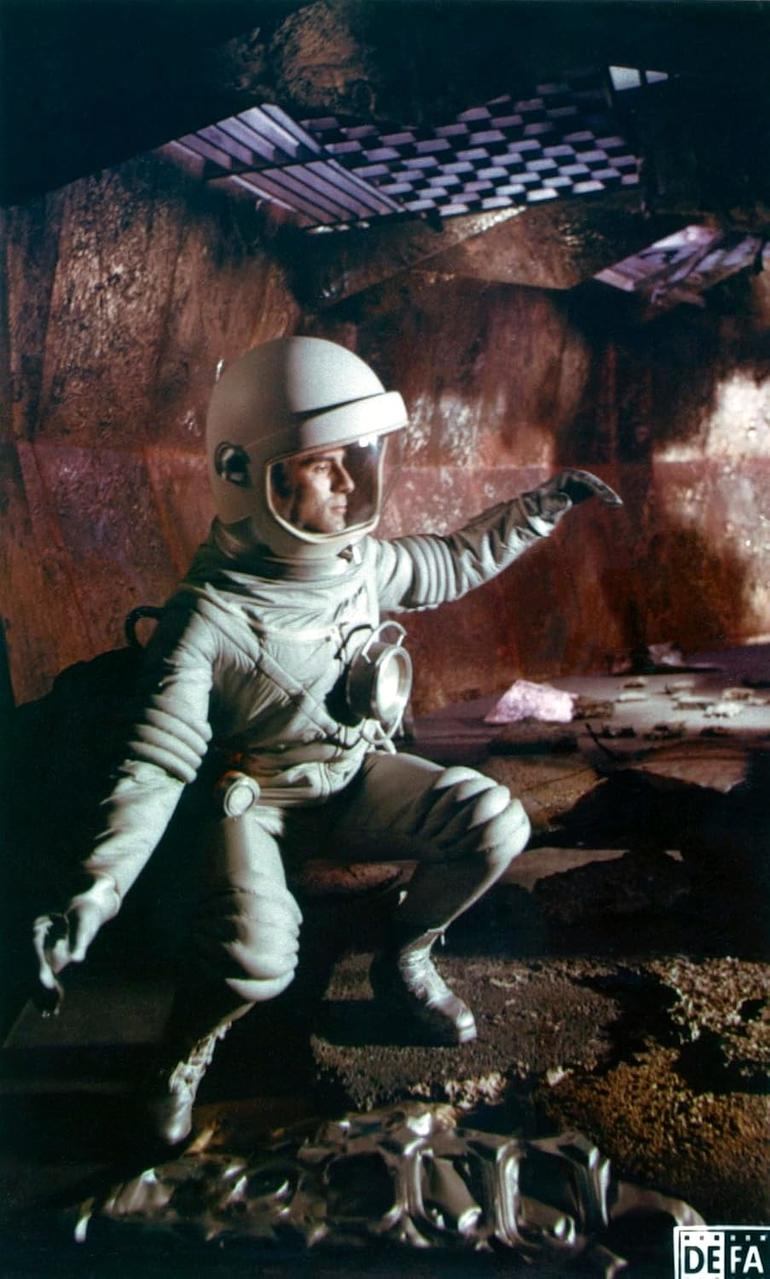 Ein Astronaut in einem weißen Raumanzug kniet in einer Höhle und zeigt mit der Hand.