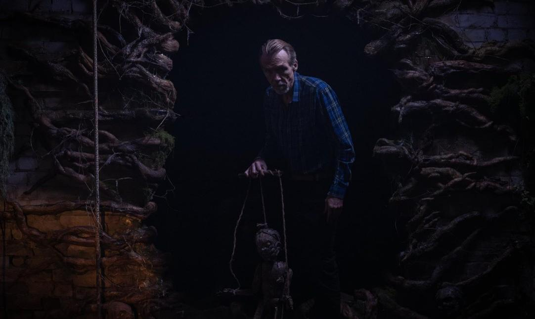 Ein Mann mit einem karierten Hemd hält eine Marionette in einer dunklen, von Wurzeln umgebenen Umgebung.