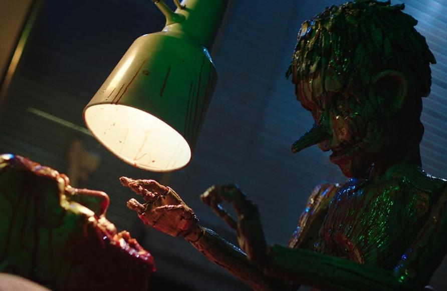 Eine Puppe mit langem Kinn blickt auf eine blutbefleckte Figur unter einer Lampe.