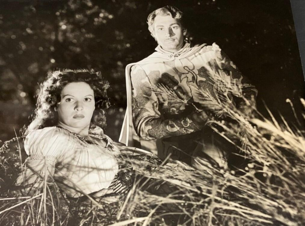 Mann mit Umhang und Frau sitzen im Gras. Dunkler Hintergrund.