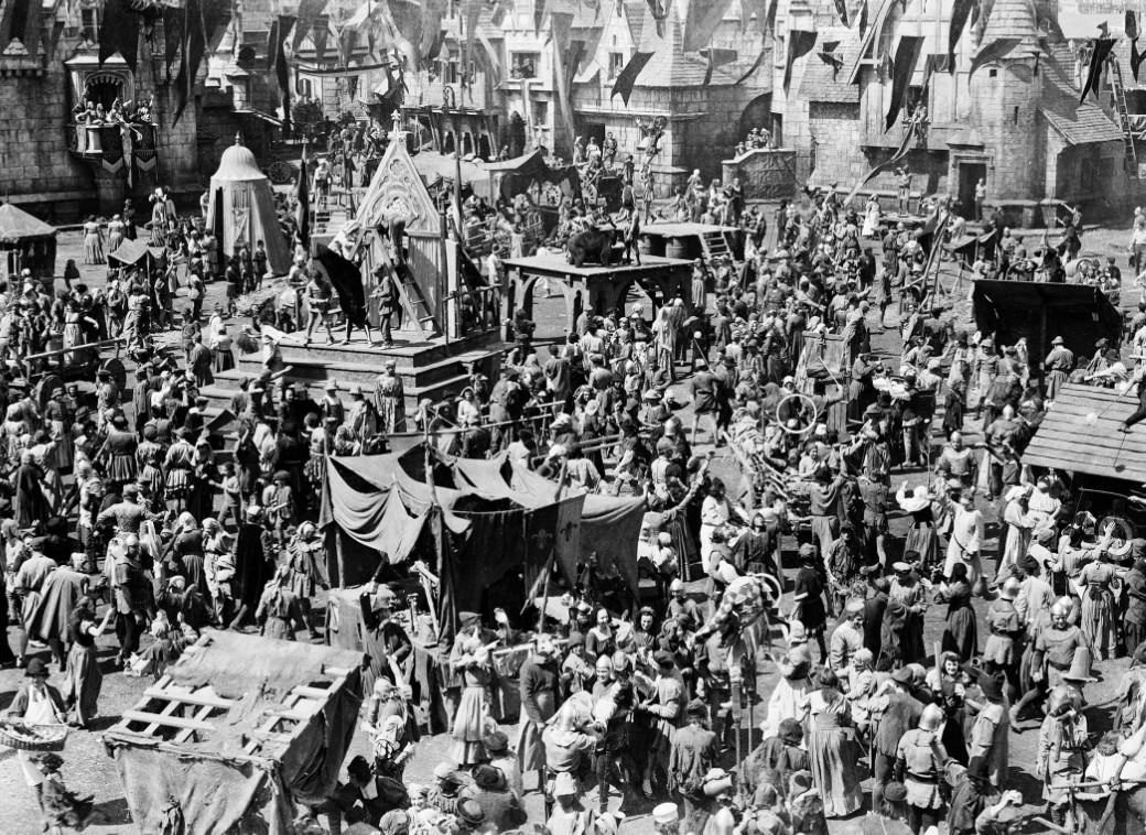 Eine mittelalterliche Stadt voller Menschen, mit Zelten und Gebäuden im Hintergrund.