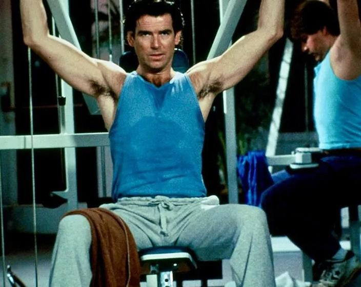 Mann trainiert im Fitnessstudio mit Gewichten, trägt blaues Tanktop und graue Hose.