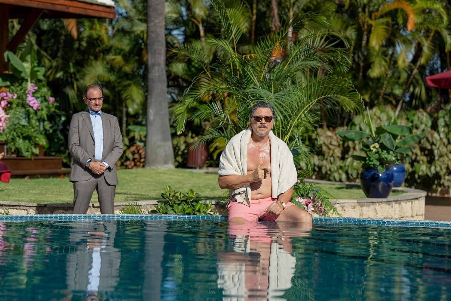 Mann sitzt am Pool mit Handtuch, Mann im Anzug steht daneben.