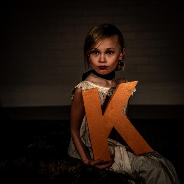 Ein junges Mädchen in einem weißen Kleid hält den orangen Buchstaben K.