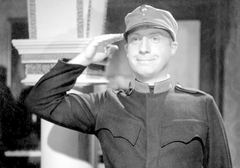 Mann in Uniform salutiert, trägt eine Mütze, lächelt leicht.