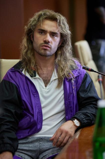 Mann mit langen blonden Haaren, lila und schwarzer Jacke, sitzt am Tisch.