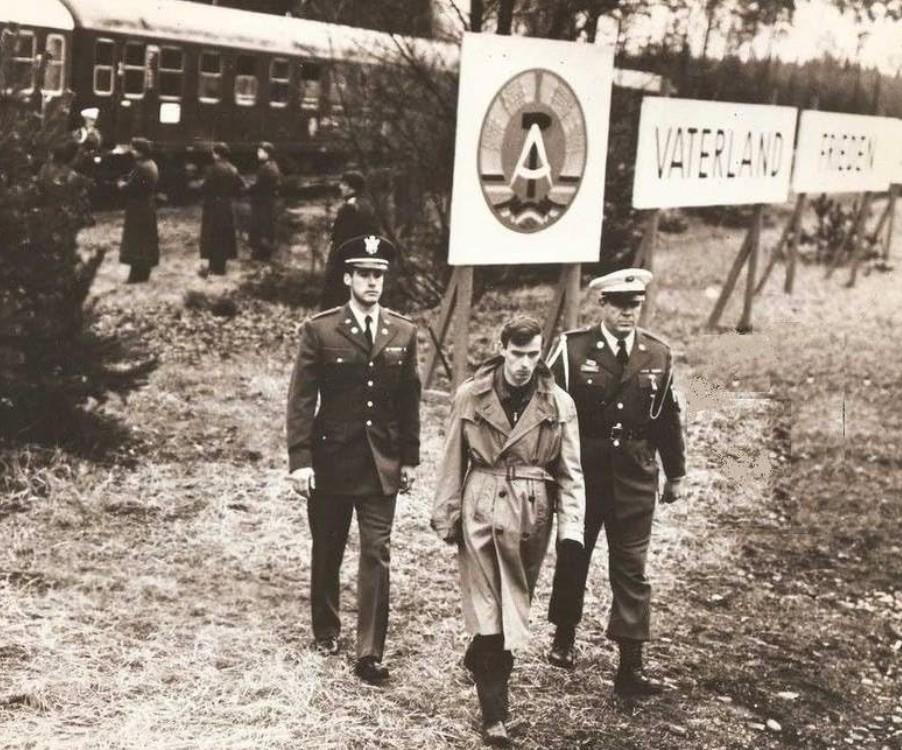 Zwei Soldaten eskortieren einen Mann mit Tarnmantel neben einem Zug und Schildern.