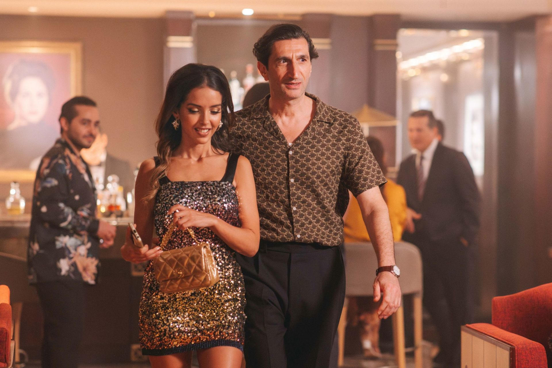 Ein Mann und eine Frau in einer Bar. Sie trägt ein glitzerndes Kleid und eine goldene Tasche. Er trägt ein gemustertes Hemd.