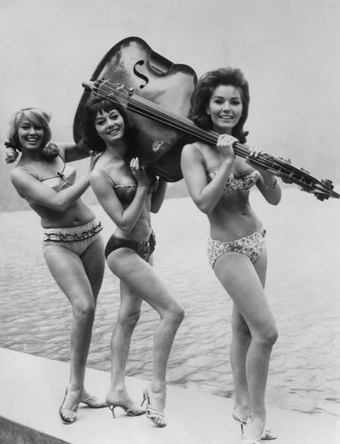 Drei Frauen in Bikinis tragen einen Kontrabass über ihren Schultern am Wasser.