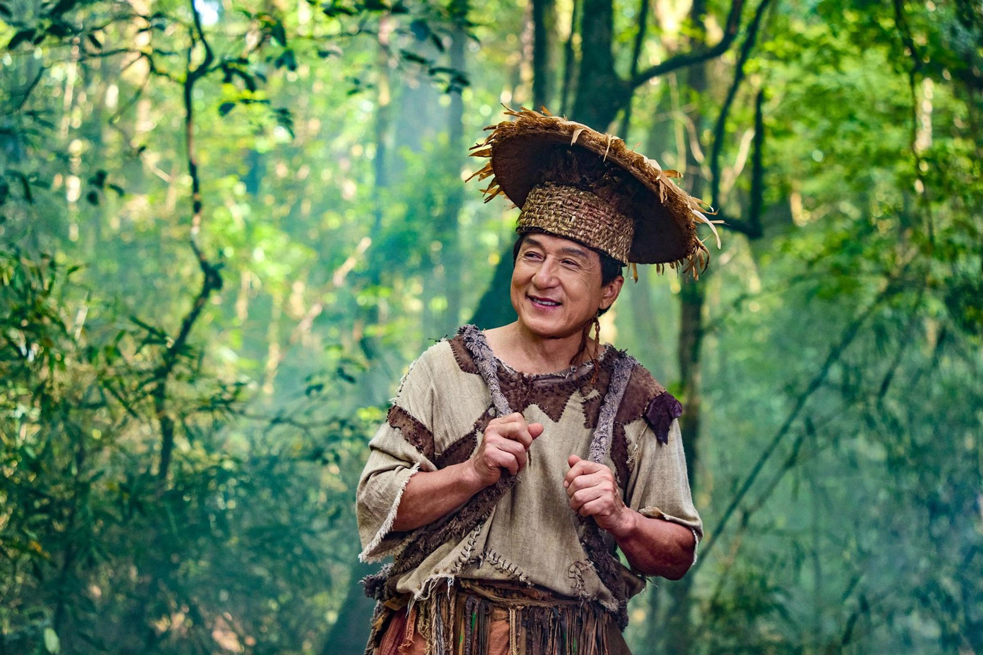 Jackie Chan in traditioneller Kleidung und einem Strohhut im Wald.