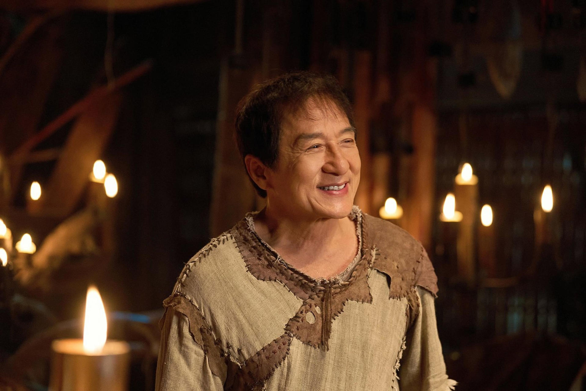 Jackie Chan lächelt in einem rustikalen Outfit, umgeben von Kerzenlicht.