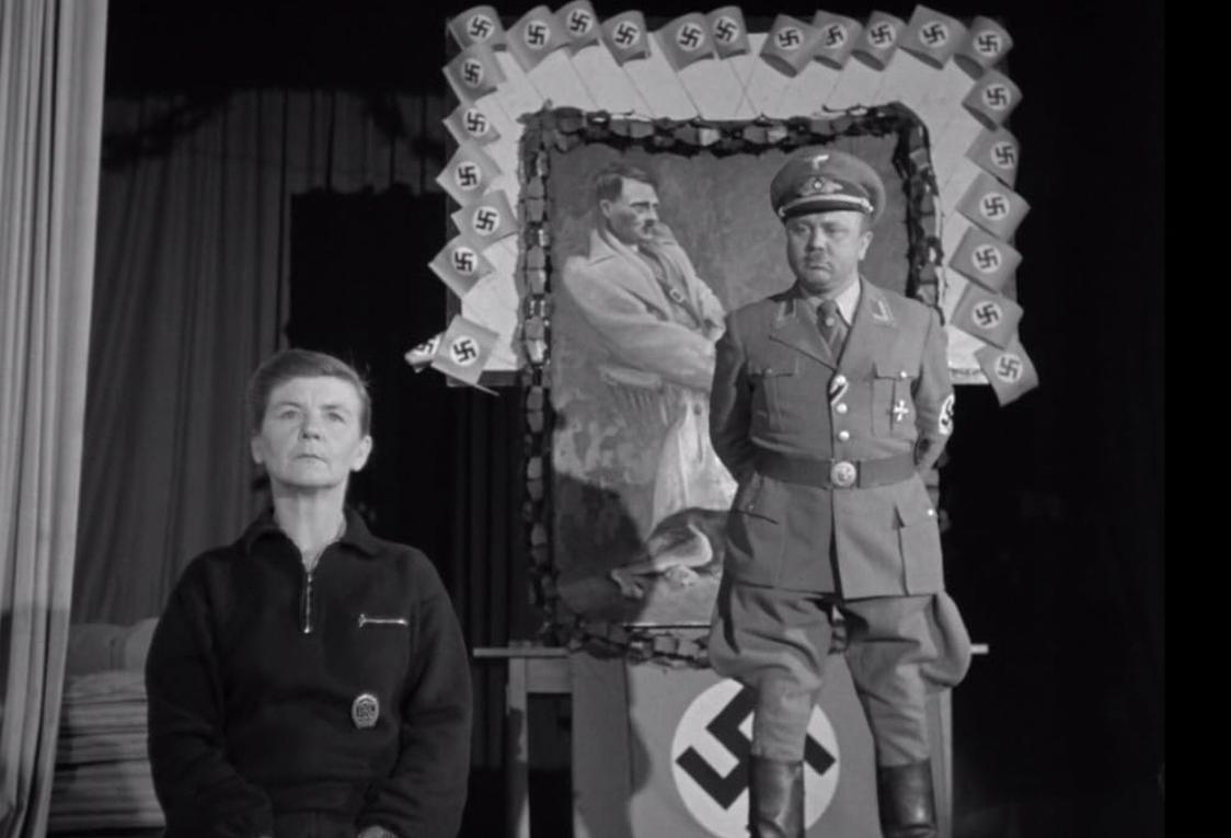 Eine Frau und ein SS-Offizier stehen vor einem Hitler-Porträt, umgeben von Hakenkreuzfahnen.
